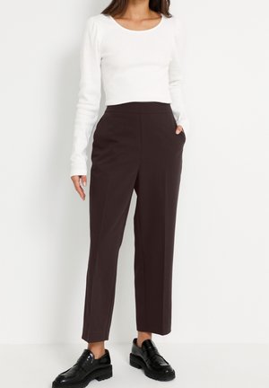 Kobieta w białej bluzce z długim rękawem, ciemnobrązowych, prostych spodniach z wysokim stanem i czarnych masywnych mokasynach, stojąca z jedną ręką w kieszeni.