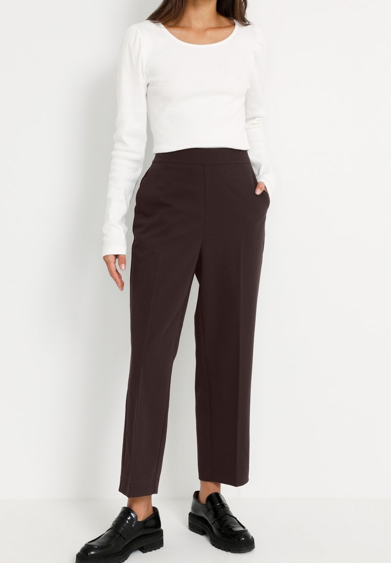 Femme portant un haut blanc à manches longues, un pantalon marron foncé taille haute, jambes droites, et des mocassins noirs épais, debout avec une main dans la poche.