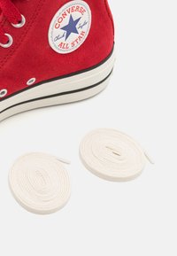 Czerwone zamszowe sneakersy w stylu high-top z białą gumową podeszwą, czarnym paskiem i naszywką z logo. Obok buta znajdują się dwie białe bawełniane sznurówki.
