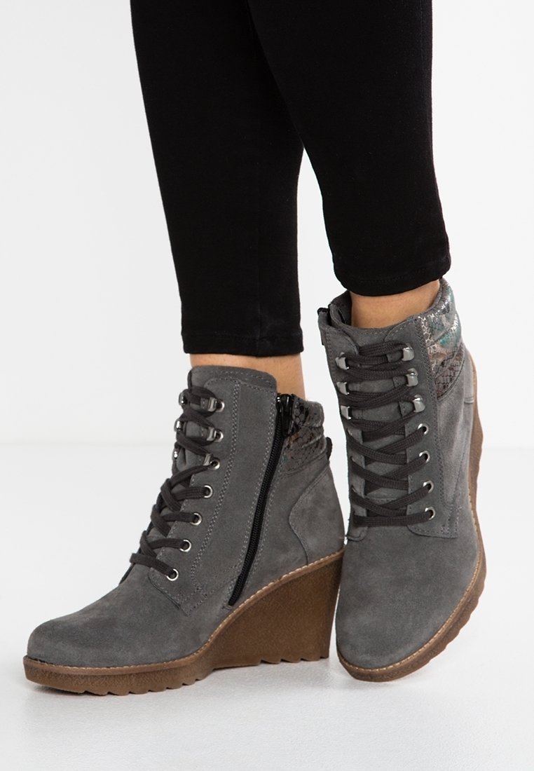 Bottes à talon compensé en daim gris avec lacets noirs, fermeture éclair latérale et col à motif, portées avec un pantalon noir.