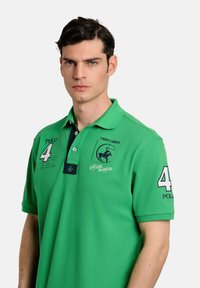 Polo verde hecho de tejido texturizado, con un collar azul marino, el número "4" blanco, logotipo bordado y acentos de texto en el pecho.