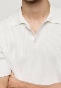 Polo blanc côtelé avec un col classique, une patte à trois boutons et des manches courtes, présentant une texture subtile et une coupe ajustée.