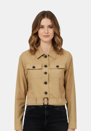 Donna che indossa una giacca beige con bottoni, tasche frontali con patta e fibbia in vita, abbinata a pantaloni neri, in piedi su sfondo bianco.