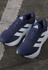 Pair of navy blue and white Adidas running shoes with laces, placed on asphalt near a faded white line.