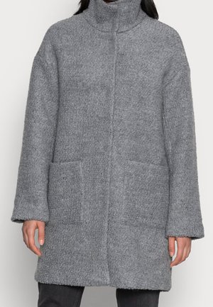 Femme portant un manteau gris texturé mi-cuisse avec un col haut et deux poches plaquées sur le devant, se tenant devant un fond uni.