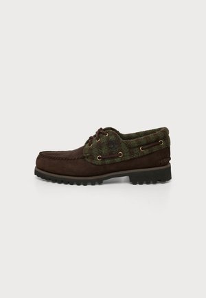 Donkerbruine leren schoen met groene geruite stoffen kraag, veterzijden en dikke zwarte rubberen zool, gezien van opzij op een witte achtergrond.
