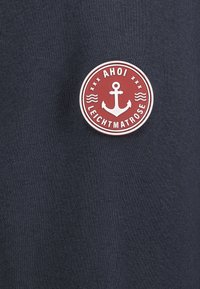 Marineblauwe stof met een ronde rode badge met een anker en de tekst "AHOI LEICHTMATROSE." Glad oppervlak, casual ontwerp.