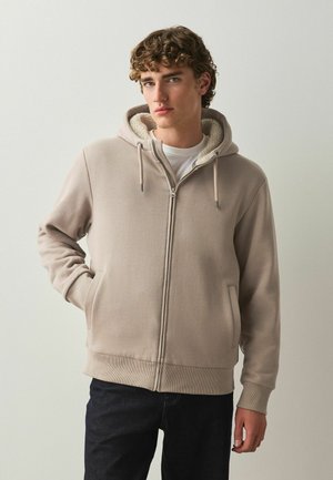 Junger Mann mit lockigem Haar, der einen beigen Hoodie mit Reißverschluss und Fleecefutter sowie dunkle Hose trägt, steht mit einer Hand in der Tasche.