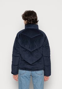 Granatowa kurtka puchowa z sztruksu z wysokim kołnierzem, z przeszyciami w kształcie szewronów na plecach i prążkowaną fakturą. Luźny fason, z długimi rękawami.