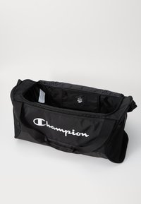 Black duffel bag made of durable fabric, featuring a prominent white "Champion" logo, zippered top, and adjustable strap. Interior has pockets.