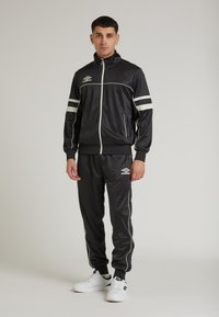 Completo sportivo nero con strisce bianche sulle maniche e sul lato dei pantaloni; realizzato in tessuto morbido; giacca con zip; logo del marchio visibile.