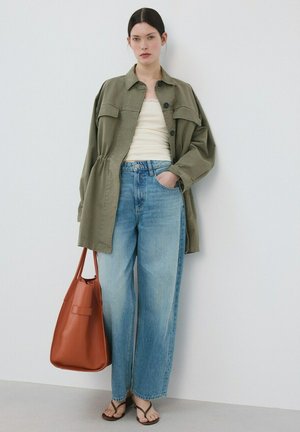 Žena stojaca opretá o stenu, má na sebe veľkú olivovú bundu, biely top, modré široké džínsy, hnedé sandále a drží veľkú hnedú koženú tašku.