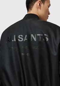 Homme portant une veste bomber noire avec le texte réfléchissant "ALLSAINTS LONDON" et une écriture cursive discrète dans le dos, la tête tournée sur le côté.