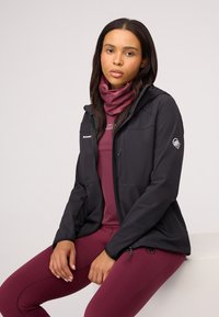 Black softshell jacket with hood, zip front, and logo on sleeve. Burgundy base layer and leggings. Smooth texture and fitted design.