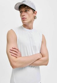 Fiatal férfi fehér ujjatlan pólót és világosszürke sapkát visel, karba tett kézzel, az oldalra néz egy egyszerű fehér háttér előtt.