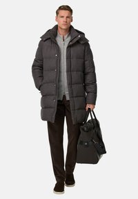 Pelēka quilted pūkains mētelis ar augstu apkakli, pelēks rāvējslēdzēja džemperis, brūni korduroja bikses un brūni sporta apavi, turot tumšu somu.