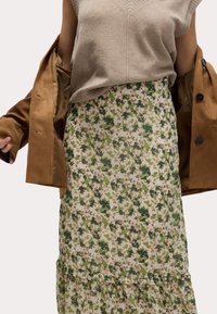 Jupe à imprimé floral crème avec des motifs de feuilles vertes, ourlet à volants superposé, associée à un top sans manches beige et une veste extérieure marron.