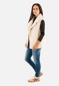 Chaleco de piel beige claro con cuello chal, superpuesto a una camiseta negra de manga larga, combinado con jeans azules y zapatillas beige con acentos rojos.