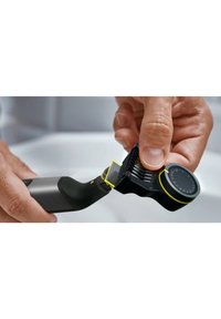 Zwarte trimmer met metalen accenten, ergonomische handgreep, displayknop en afneembare beschermkap, met gele accenten en een gestructureerde grip.