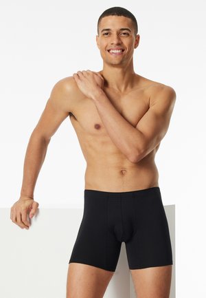 Schwarze Baumwoll-Boxershorts mit eng anliegendem Schnitt, ausgestattet mit einer glatten Textur und einem klassischen Bund, präsentiert von einer Person vor neutralem Hintergrund.