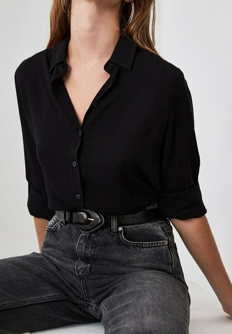 Camicia nera con bottoni e maniche arrotolate, realizzata in tessuto morbido, abbinata a jeans grigio scuro a vita alta e cintura nera.