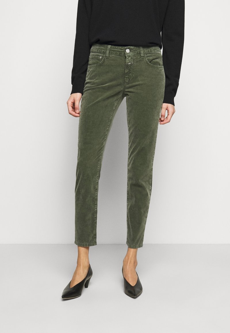 Femme portant un pantalon slim en velours côtelé vert olive associé à un haut noir à manches longues et des chaussures noires à talons bas et bouts pointus, debout.