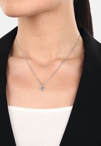 Silverhalsband med ett bladformat hänge, prytt med en klunga av små stenar, fäst vid en tunn kedja. Bärs över en vit topp.