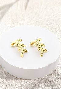 Örhängen i form av guldblad med utskurna mönster, som har en glänsande yta och knappback, presenterade på en slät vit, rund bas.