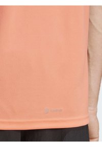 Camiseta atlética de color naranja claro con un tejido texturizado y transpirable, que presenta un dobladillo redondeado y el logo "AEROREADY" cerca del borde inferior.