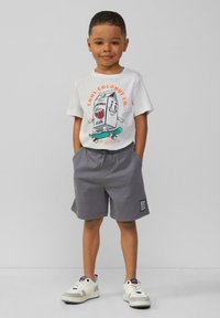 Witte t-shirt met een kleurrafische afbeelding van een karton dat op een skateboard rijdt; grijze shorts; witte sneakers met zwarte en rode accenten.