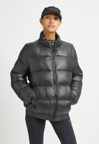 Giacca a vento nera con colletto alto, chiusura con zip e forma quadrata. Tessuto lucido e liscio con cuciture orizzontali e tasche laterali.