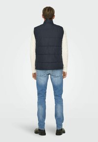 Gilet rembourré bleu marine avec col haut, porté par-dessus un pull en maille torsadée crème. Associe à un jean bleu clair et des chaussures noires.