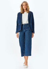 Chaqueta de denim con cuello muesca y dos bolsillos frontales, combinada con jeans de pierna ancha de tiro alto. Ambos en azul oscuro, estética casual.