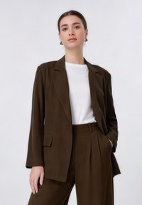 Femme aux cheveux foncés portant un blazer marron et un pantalon assorti, posant devant un fond blanc uni, vêtue d'une chemise blanche et portant des boucles d'oreilles.