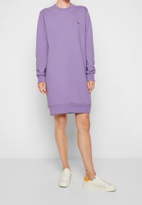 Žena v dlouhém rukávu, šatích po kolena levandulové barvy s malým zvířecím logem, v kombinaci s bílými teniskami se žlutými akcenty.