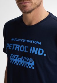 T-maica iz temno modrega bombaža z bold modrim napisom: "NASCAR CUP DAYTONA," "PETROL IND.," in "EST. 1989" z izstopajočim šahiranim vzorcem.