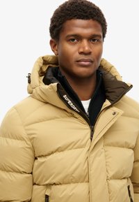 Chaqueta de plumas beige con capucha, que presenta un diseño bicolor, cierre con cremallera y acentos de marca. El material parece suave y aislante.