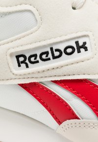 Zapatillas de cuero blanco y sintético con detalles en rojo y el logo característico de Reebok. Superficie texturizada con costuras decorativas.