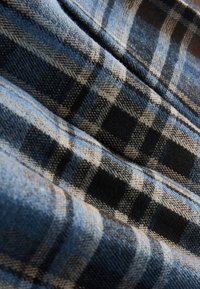 Niebiesko-czarna tkanina w kratę z beżowymi akcentami; posiada teksturowaną powierzchnię i widoczne włókna, złożona z falami tworzącymi głębię.