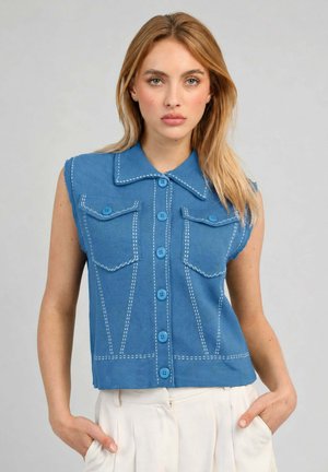 Femme aux longs cheveux blonds portant une chemise bleue sans manches boutonnée avec des coutures blanches et un pantalon blanc, debout les mains dans les poches.