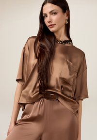 Top in satin marrone con maniche corte, dettaglio tasca e vestibilità rilassata, abbinato a pantaloni coordinati e una collana d'oro chunky.