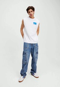 White sleeveless t-shirt with a blue logo, paired with blue cargo jeans featuring multiple pockets and contrasting stitching, completed with white sneakers.