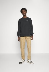 Felpa nera con logo "GANT 1948", pantaloni beige e sneakers in bianco e nero. Design semplice, collo a giro e polsini a costine.