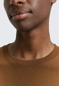 Bruine t-shirt met ronde hals en een gladde textuur, met een getailleerd ontwerp en een platte geribde halslijn. Huidskleur zichtbaar onder de kraag.