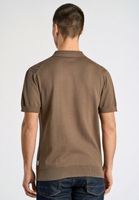 Mies, jolla on lyhyet hiukset, pukeutuneena ruskeaan lyhythihaiseen neulottuun polo-paitaan ja tummansiniin farkkuihin, kuvattuna selkäpuolelta yksinkertaista taustaa vasten.