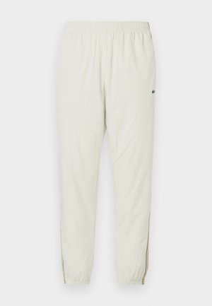 Jogging léger crème avec une taille élastique, des chevilles resserrées, des fentes latérales et un petit logo vert sur la cuisse supérieure.