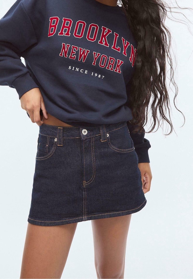 Personne portant un sweat-shirt bleu marine avec le texte "Brooklyn New York Since 1987" et une mini-jupe en jean bleu foncé avec des poches et des détails de coutures.