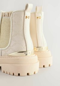 Weiße Stiefeletten aus strukturiertem Leder, mit goldenen Akzenten und einer klobigen Sohle. Sie verfügen über ein gemustertes Design und elastische Seitenpaneele.