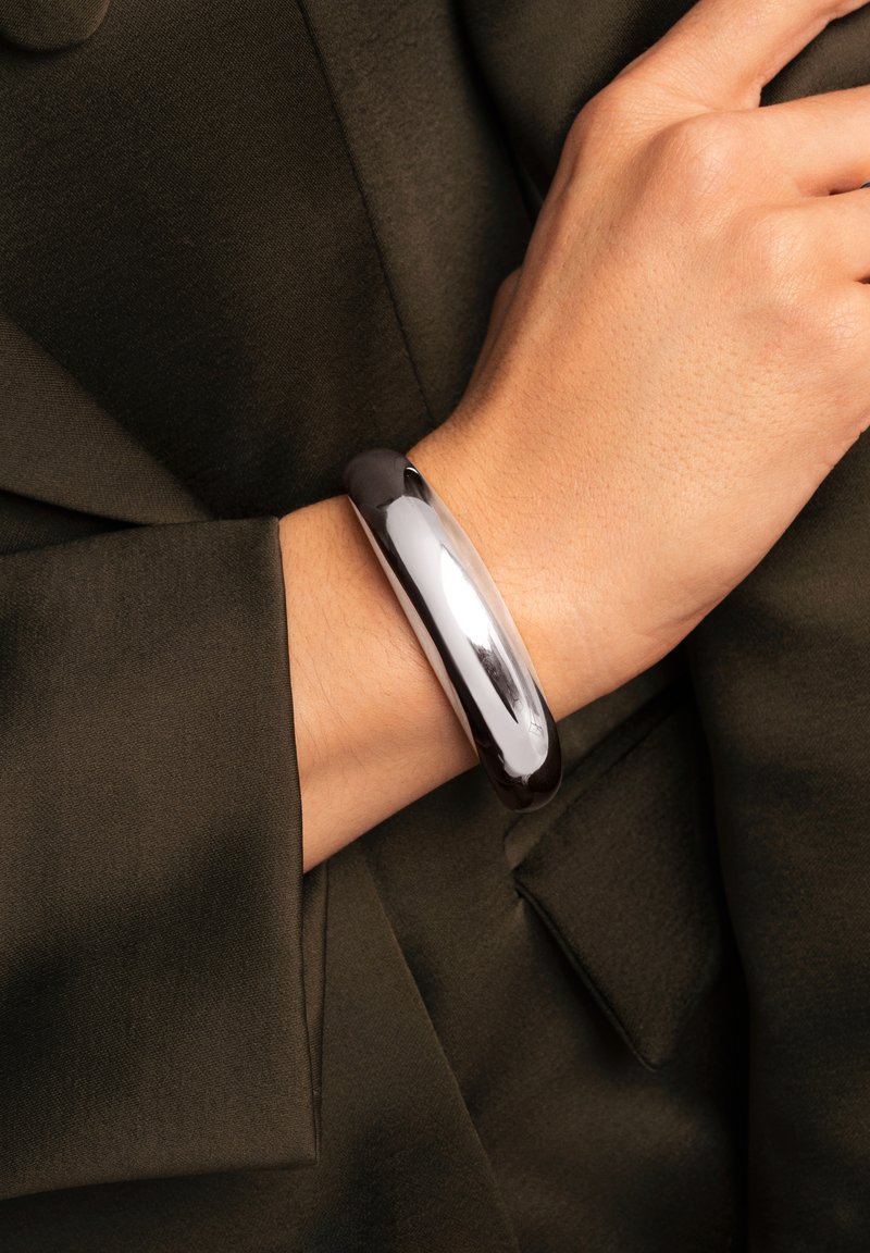 Metallarmband mit glatter, reflektierender Oberfläche in Silber, getragen am Handgelenk, kombiniert mit einem olivgrünen Stoffkleidungsstück.