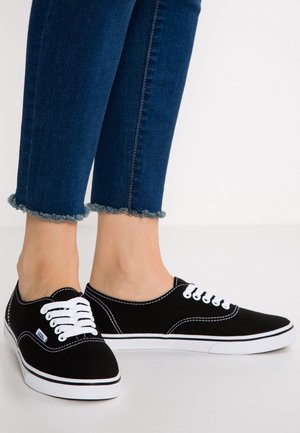 Zwarte canvas sneakers met witte veters en stiksel, voorzien van een afgeronde neus en witte rubberen zool. Jeans met uiteengerafelde zoom zichtbaar.